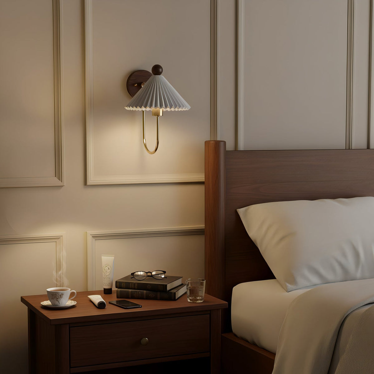 A Clérachic wall lamp with a pleated shade lit above a dark wood bedside table with a teacup, books and glasses, by a bed with cream pillows.