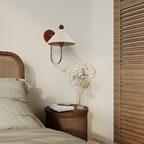 This Clérachic wall lamp has a pleated white shade above a rattan bedside table, beside a cane-headed bed and a vase of dried flowers.