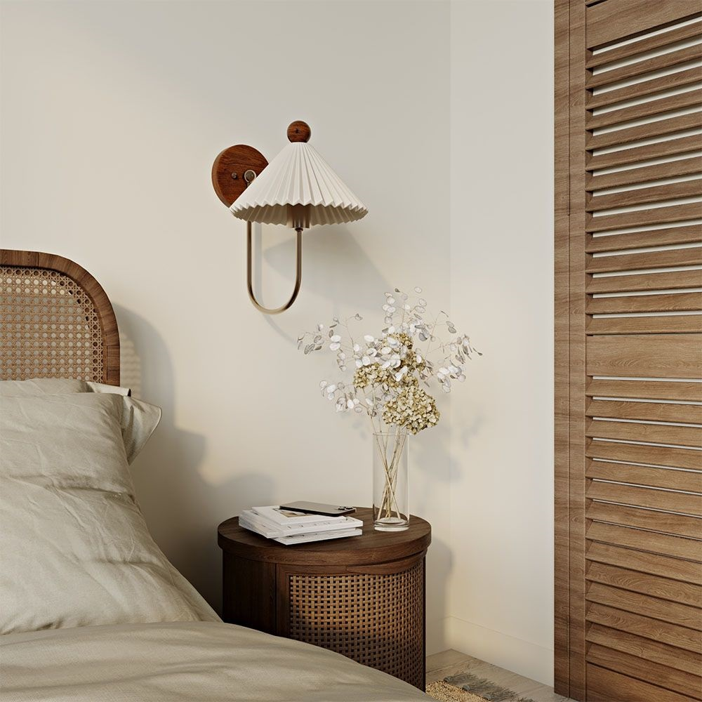 This Clérachic wall lamp has a pleated white shade above a rattan bedside table, beside a cane-headed bed and a vase of dried flowers.