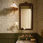 Shown here, a Clérachic wall lamp with a pleated shade beside an ornate dark mirror, against green leafy wallpaper above a basin with brass taps.