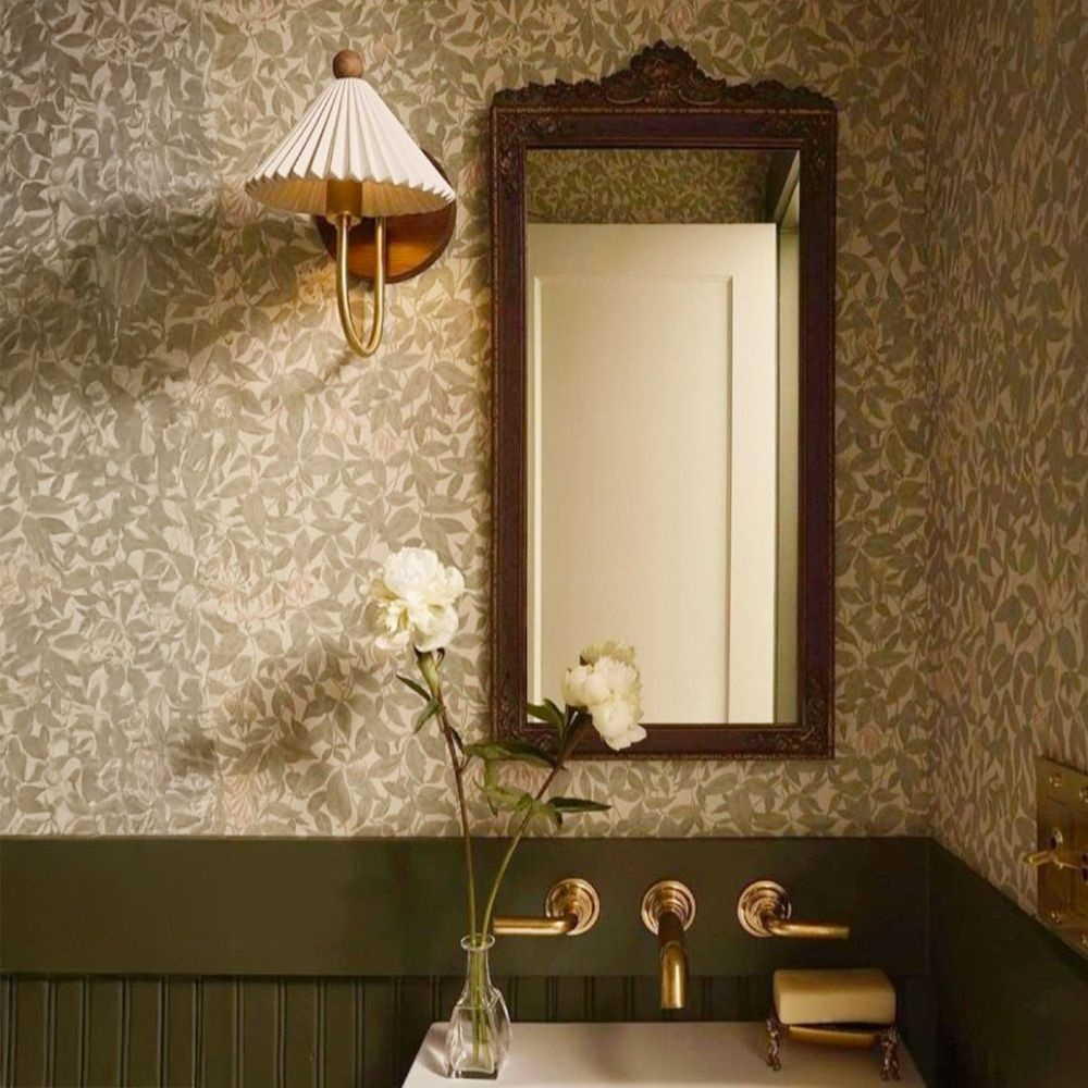 Shown here, a Clérachic wall lamp with a pleated shade beside an ornate dark mirror, against green leafy wallpaper above a basin with brass taps.