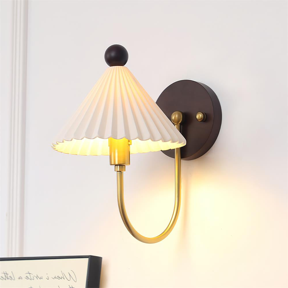 Close view of a lit Clérachic wall lamp, with a pleated white shade, a brass fitting and a round walnut backplate on a pale wall.
