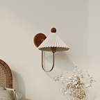 A Clérachic wall lamp with a pleated white shade and slim brass arm on a pale wall, with a cane bedhead and dried flowers below.