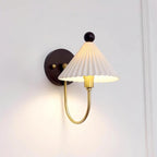 A lit Clérachic wall lamp with a pleated white shade glowing warm on a pale wall, with a curved brass arm and a round dark backplate.