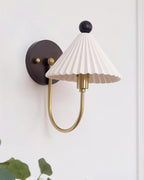 This Clérachic wall lamp has a pleated white shade and a curved brass arm on a pale panelled wall, with a green leaf at the bottom edge.