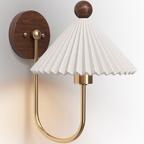 Close view of a Clérachic wall lamp lit warm, showing the pleated white shade, the brass arm and the round walnut-effect backplate.