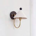 A Clérachic wall lamp with a pleated white shade, a short brass arm and a round dark backplate on a plain white wall.