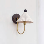 A Clérachic wall lamp with a pleated white shade, a short brass arm and a round dark backplate on a plain white wall.