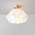 The Clérachic Pleated Petal Ceiling Light seen at an angle in a grey studio, its brass canopy holding the ruffled acrylic shade.