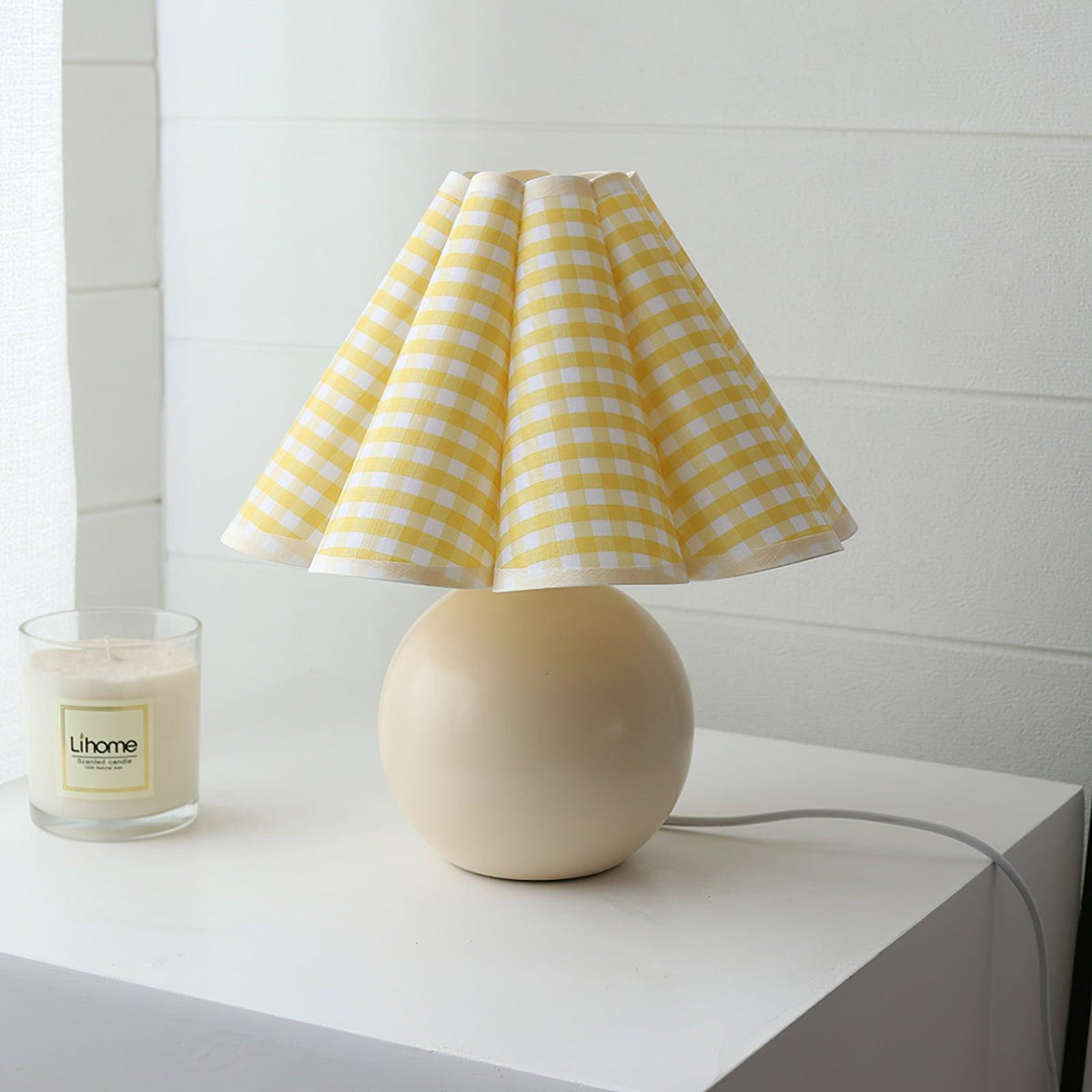 This Clérachic table lamp, with a yellow plaid pleated shade on a round beige base, stands on a white shelf beside a candle.