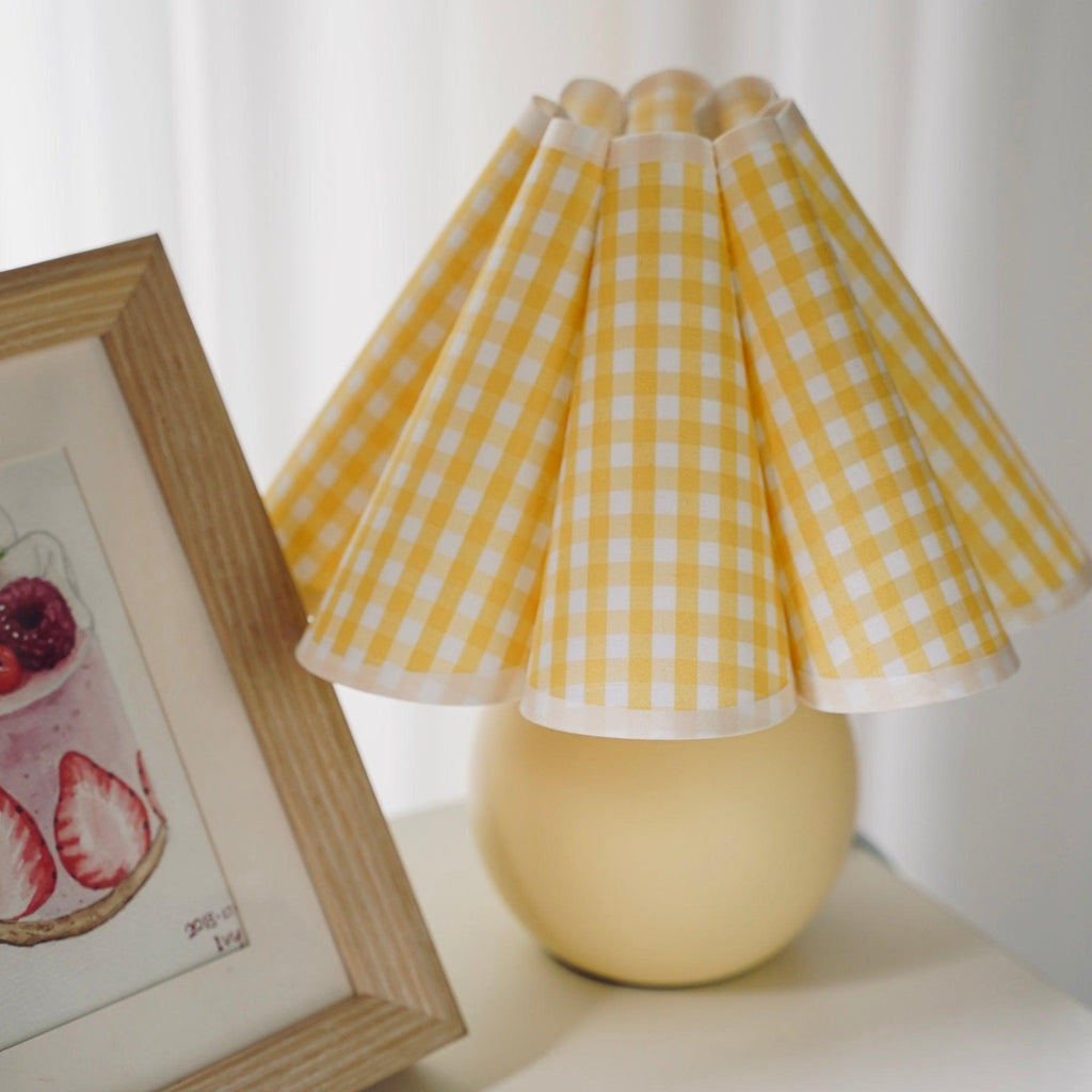 Close view of the yellow plaid pleated shade on this Clérachic table lamp, with a framed strawberry print leaning behind it.