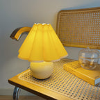 Lit up, this Clérachic table lamp with a solid yellow pleated shade glows on a rattan table beside a glass jar and a yellow book.