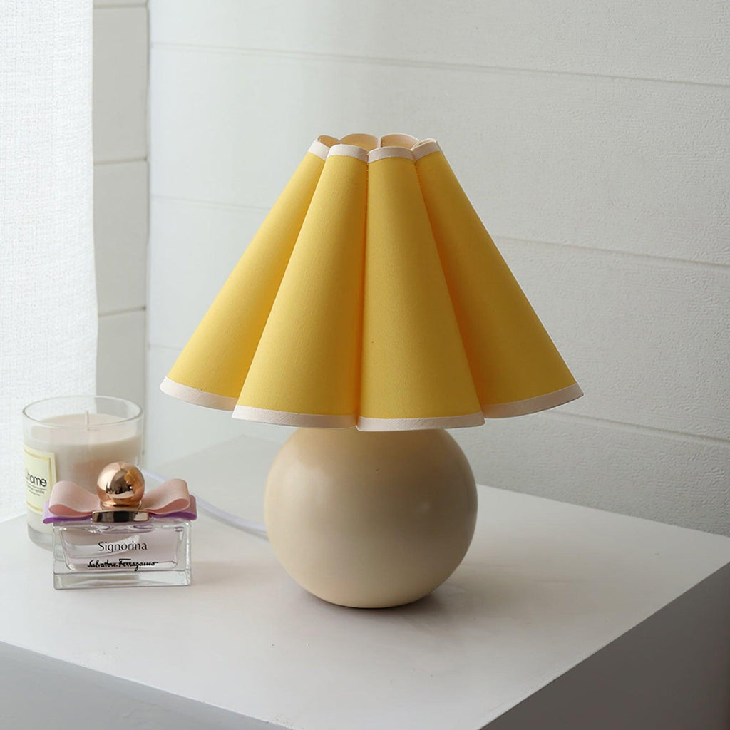This Clérachic table lamp with a solid yellow pleated shade and beige ceramic ball base stands on a white shelf next to a perfume bottle.