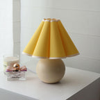 This Clérachic table lamp with a solid yellow pleated shade and beige ceramic ball base stands on a white shelf next to a perfume bottle.