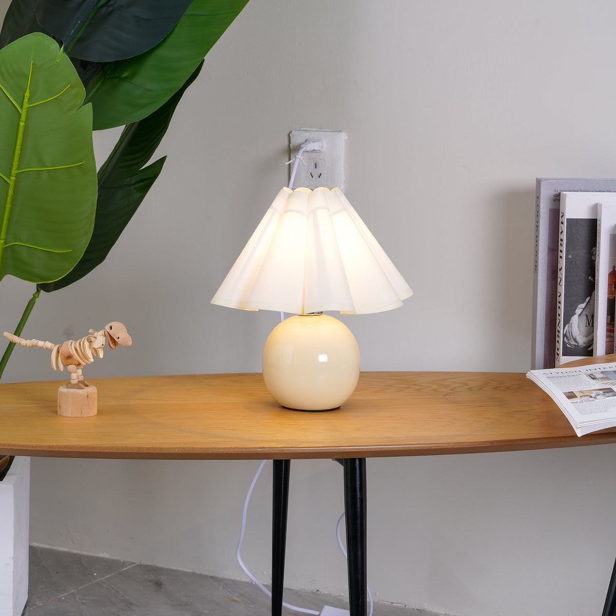 In this photo, a Clérachic table lamp with a white pleated shade glows on an oval wooden table, beside a wooden toy dinosaur and a large leaf.