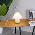 Seen a little closer, this Clérachic table lamp with a white pleated shade lights an oval wooden table next to a stack of books.