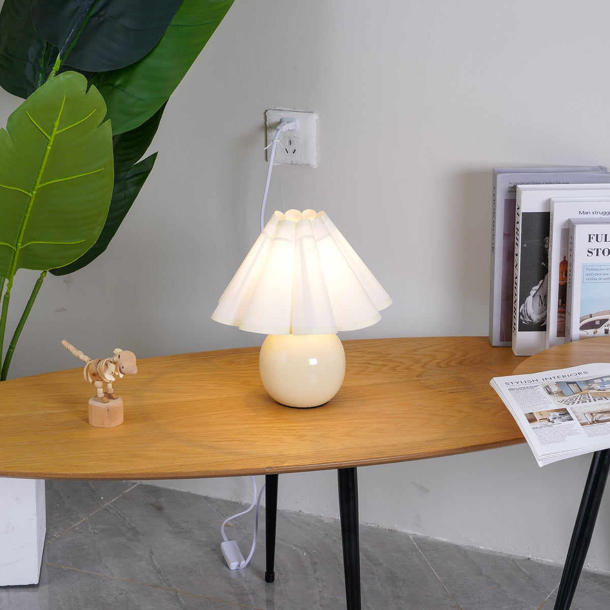 Seen a little closer, this Clérachic table lamp with a white pleated shade lights an oval wooden table next to a stack of books.