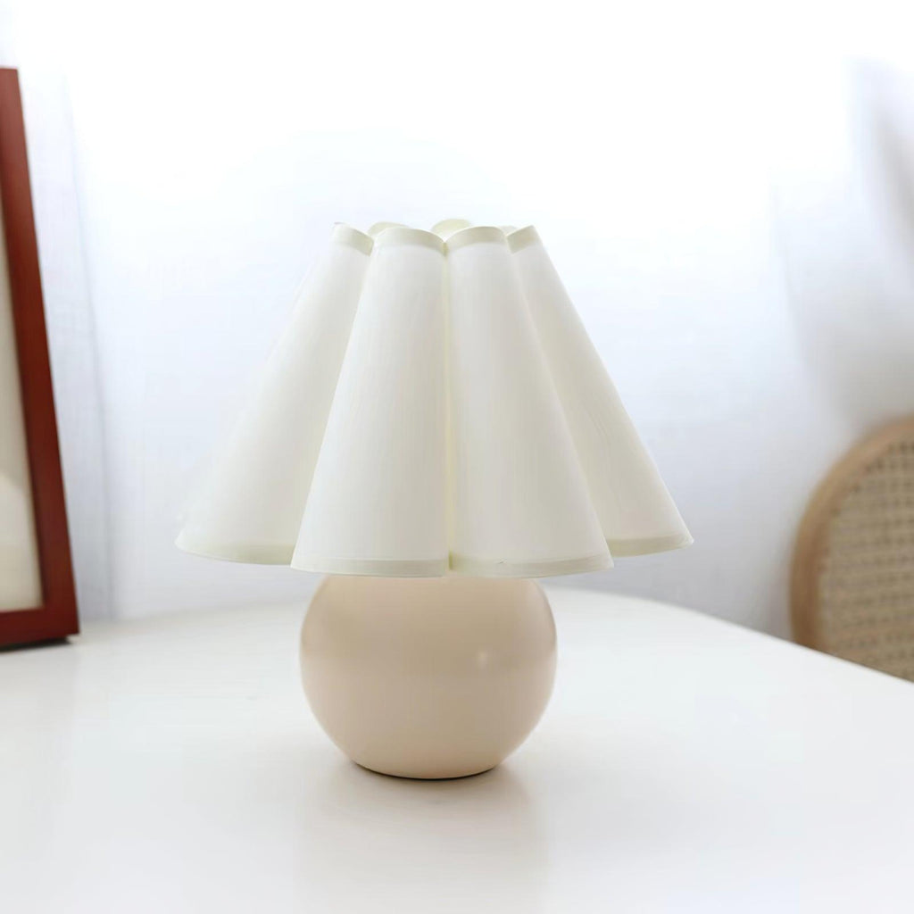 Pictured here, a Clérachic table lamp with a white pleated shade and a cream ceramic ball base on a white table by a window.