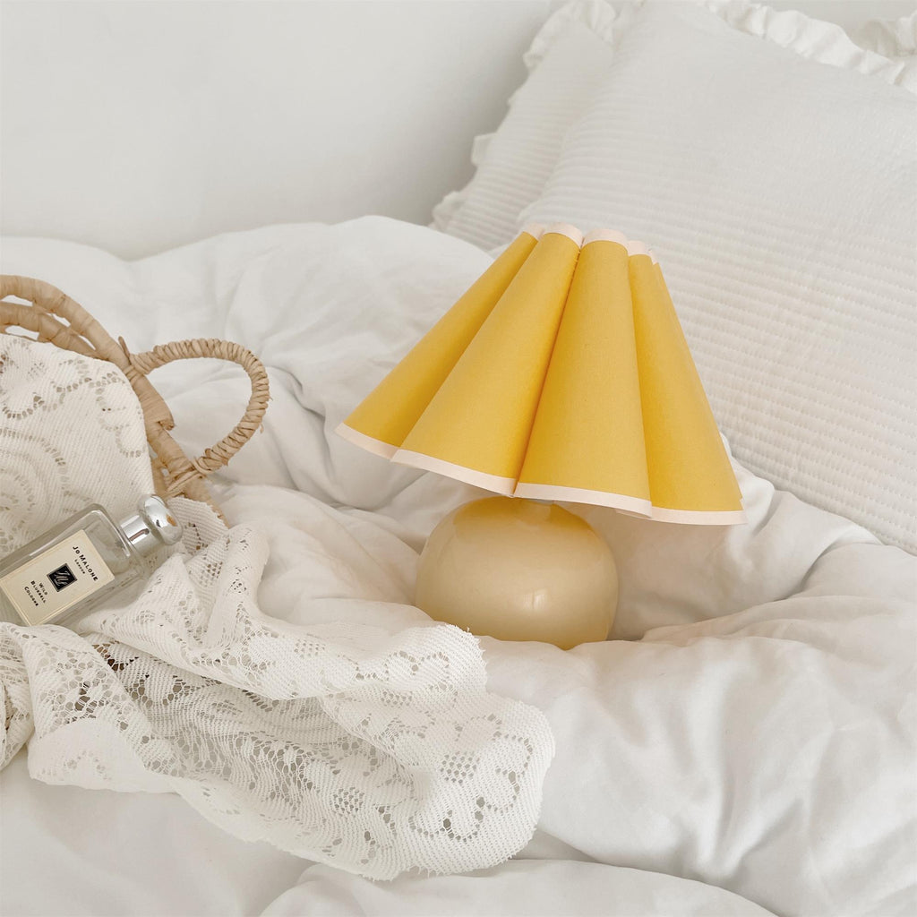 This Clérachic table lamp with a yellow pleated shade rests on a white duvet, beside lace and a small woven bag.