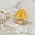 This Clérachic table lamp with a yellow pleated shade rests on a white duvet, beside lace and a small woven bag.