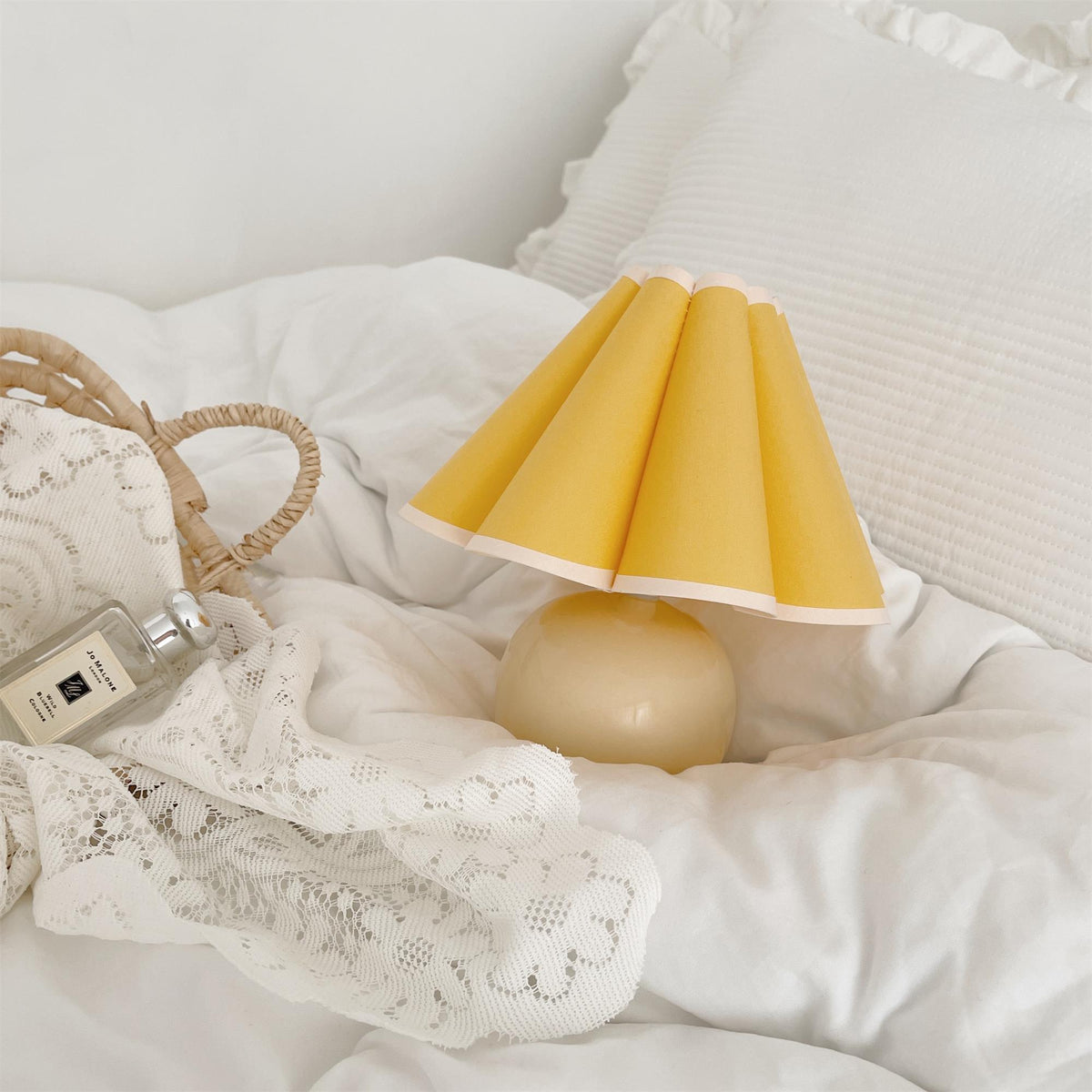 This Clérachic table lamp with a yellow pleated shade rests on a white duvet, beside lace and a small woven bag.