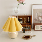 A Clérachic table lamp with a solid yellow pleated shade stands on a white dressing table, beside a hand mirror and a small wooden shelf.
