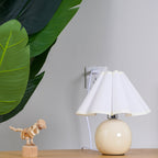This Clérachic table lamp with a white pleated shade stands on a wooden shelf, plugged into the wall beside a leaf and a toy dinosaur.