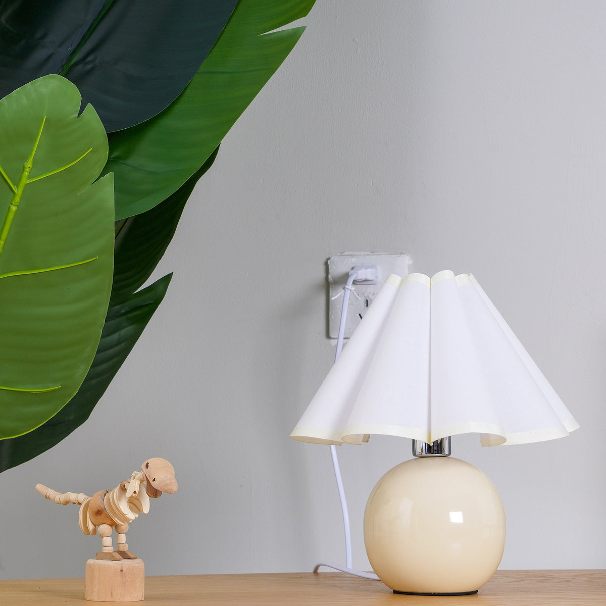 This Clérachic table lamp with a white pleated shade stands on a wooden shelf, plugged into the wall beside a leaf and a toy dinosaur.