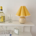 In this photo, a Clérachic table lamp with a yellow pleated shade stands on a white wall shelf, next to a wine bottle and a glass.
