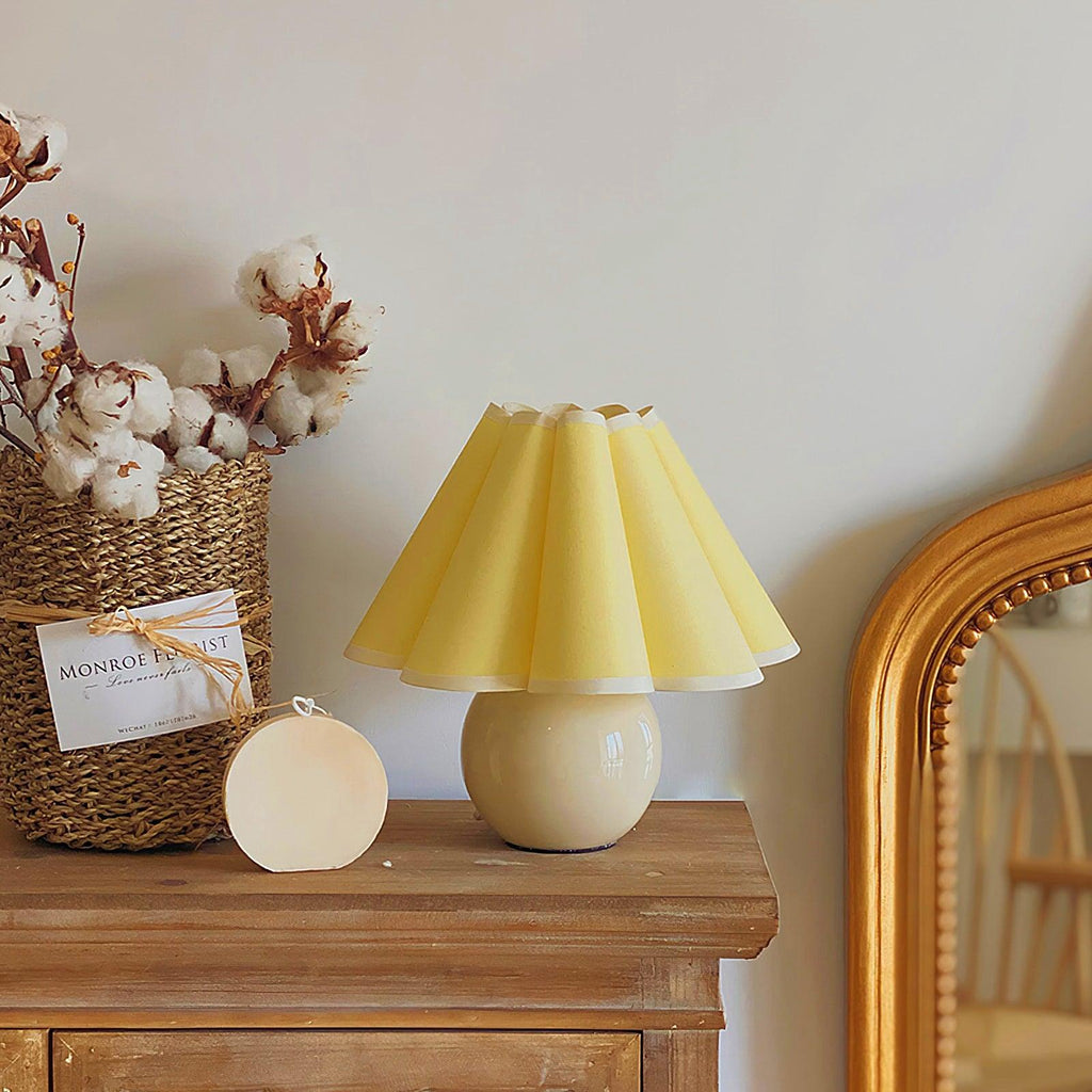 This Clérachic table lamp with a pale yellow pleated shade sits on a wooden chest, beside a basket of cotton stems and a gold mirror.