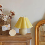 This Clérachic table lamp with a pale yellow pleated shade sits on a wooden chest, beside a basket of cotton stems and a gold mirror.