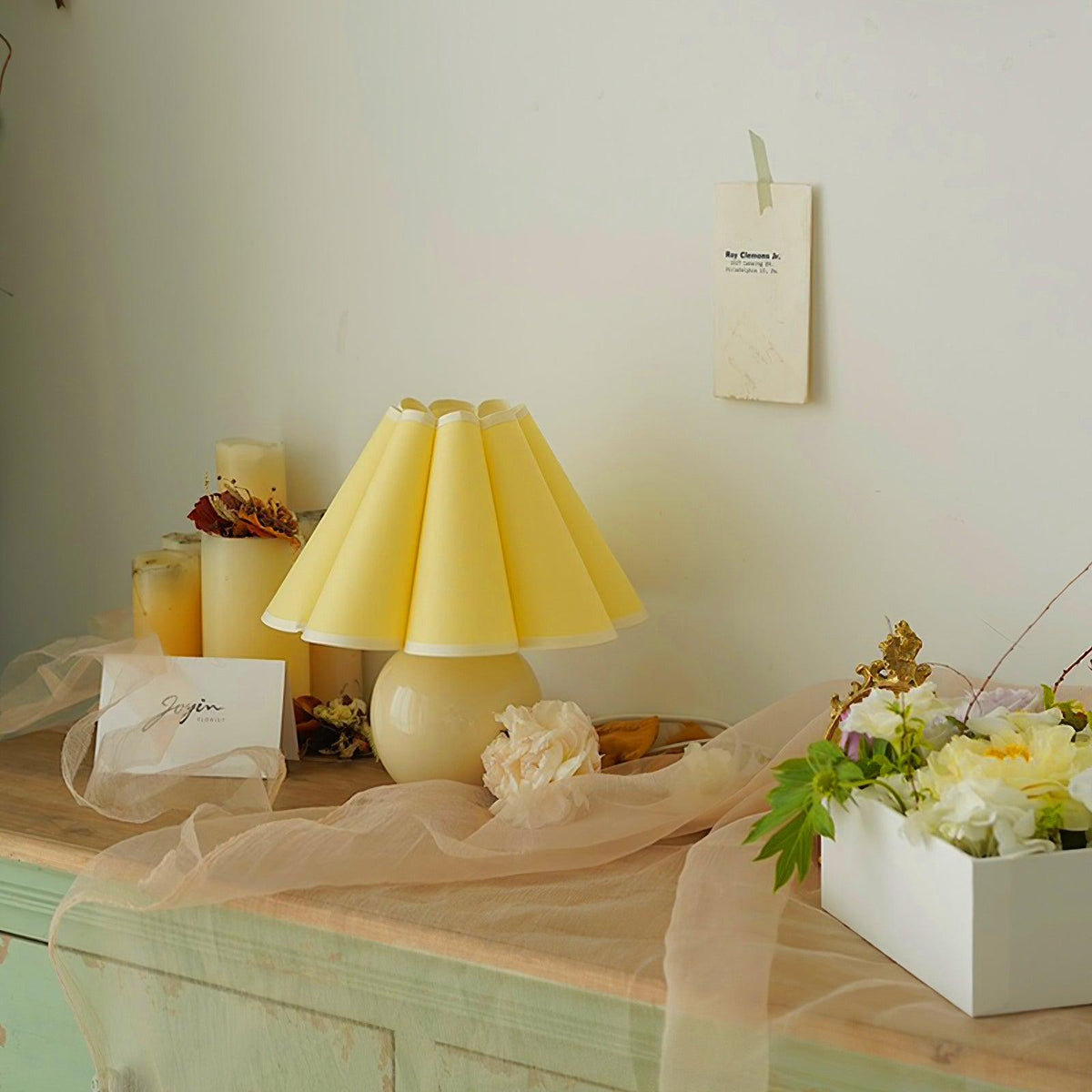 Pictured here, a Clérachic table lamp with a pale yellow pleated shade stands on a mint-green cabinet, with flowers and candles around it.