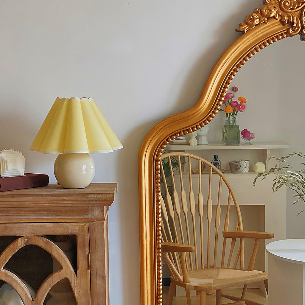 A pale yellow pleated Clérachic table lamp stands on a wooden cabinet beside a large gold-framed mirror and a wooden chair.