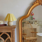 A pale yellow pleated Clérachic table lamp stands on a wooden cabinet beside a large gold-framed mirror and a wooden chair.