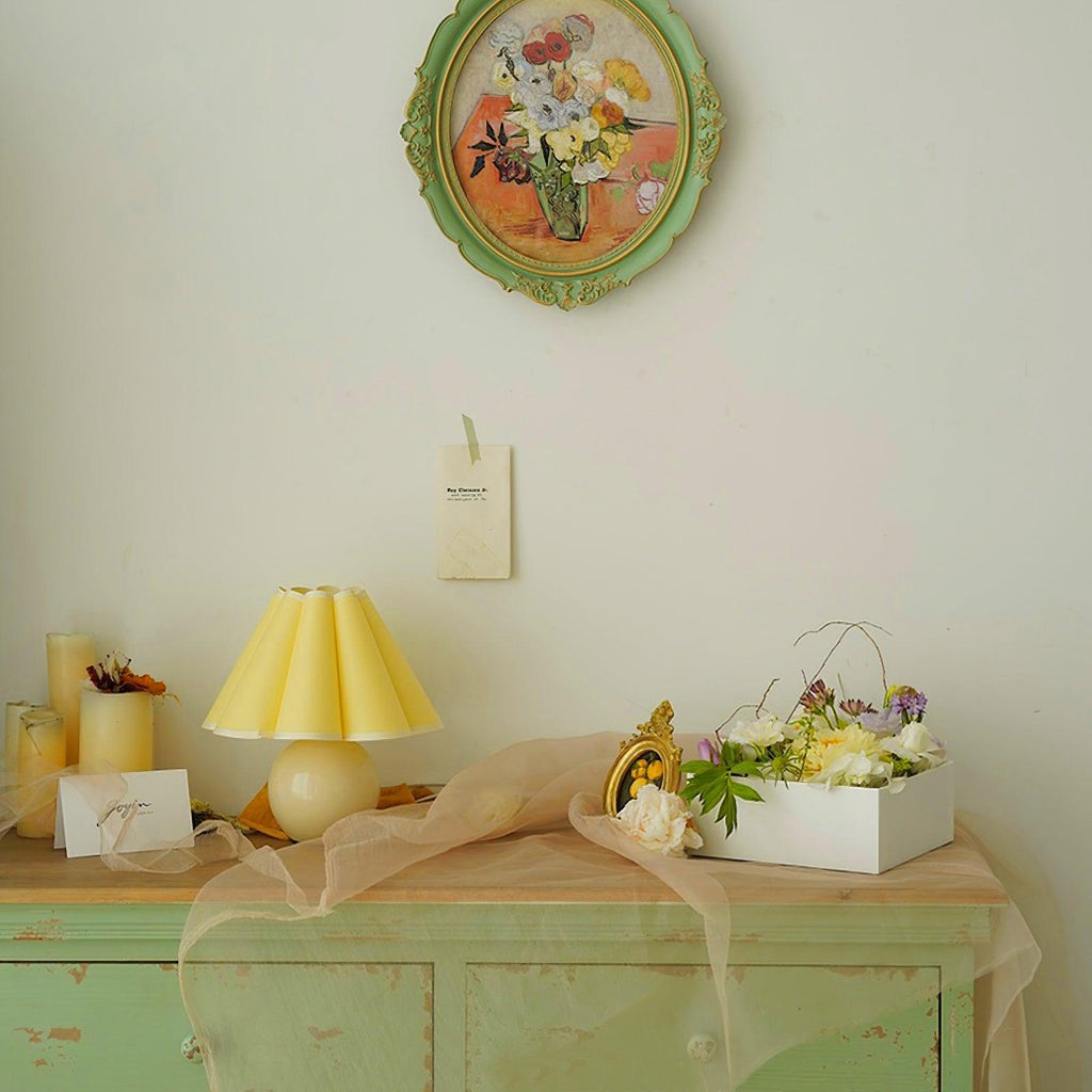 Shown here, a Clérachic table lamp with a yellow pleated shade on a mint-green cabinet, below a green oval frame and a gauze drape.