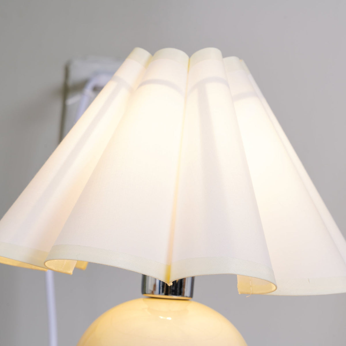 Close view of the white pleated shade on this Clérachic table lamp, lit from within with a chrome fitting below.