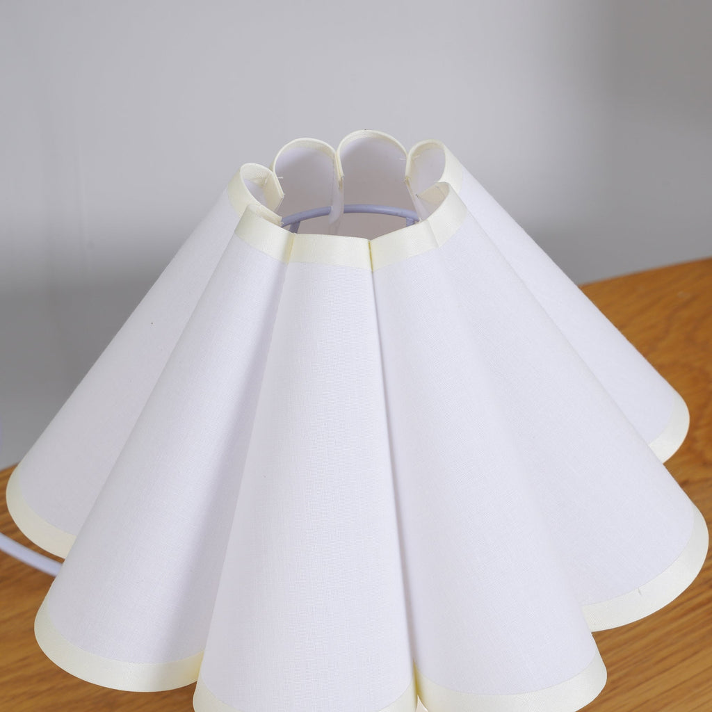 Close view of the scalloped top edge and white pleats of the shade of this Clérachic table lamp, on a wooden surface.