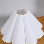 Close view of the scalloped top edge and white pleats of the shade of this Clérachic table lamp, on a wooden surface.