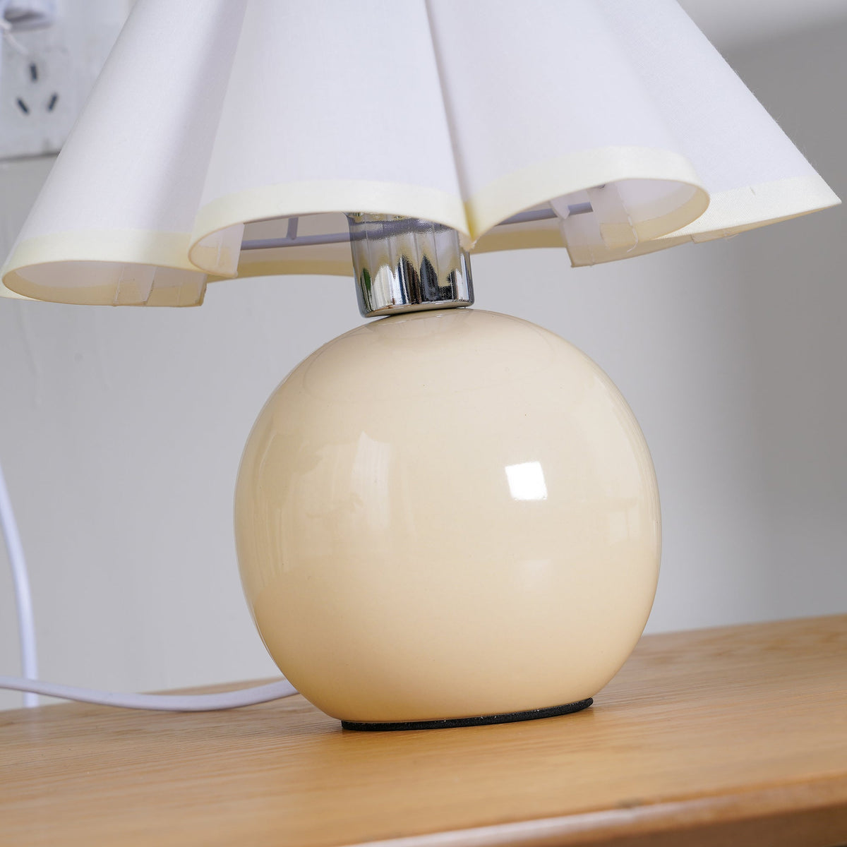 Close view of the glossy beige ceramic ball base and chrome neck of this Clérachic table lamp, under a white pleated shade.