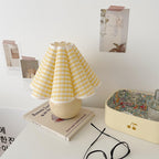 This Clérachic table lamp with a yellow plaid pleated shade stands on a white desk, with a book, glasses and pinned photos behind.