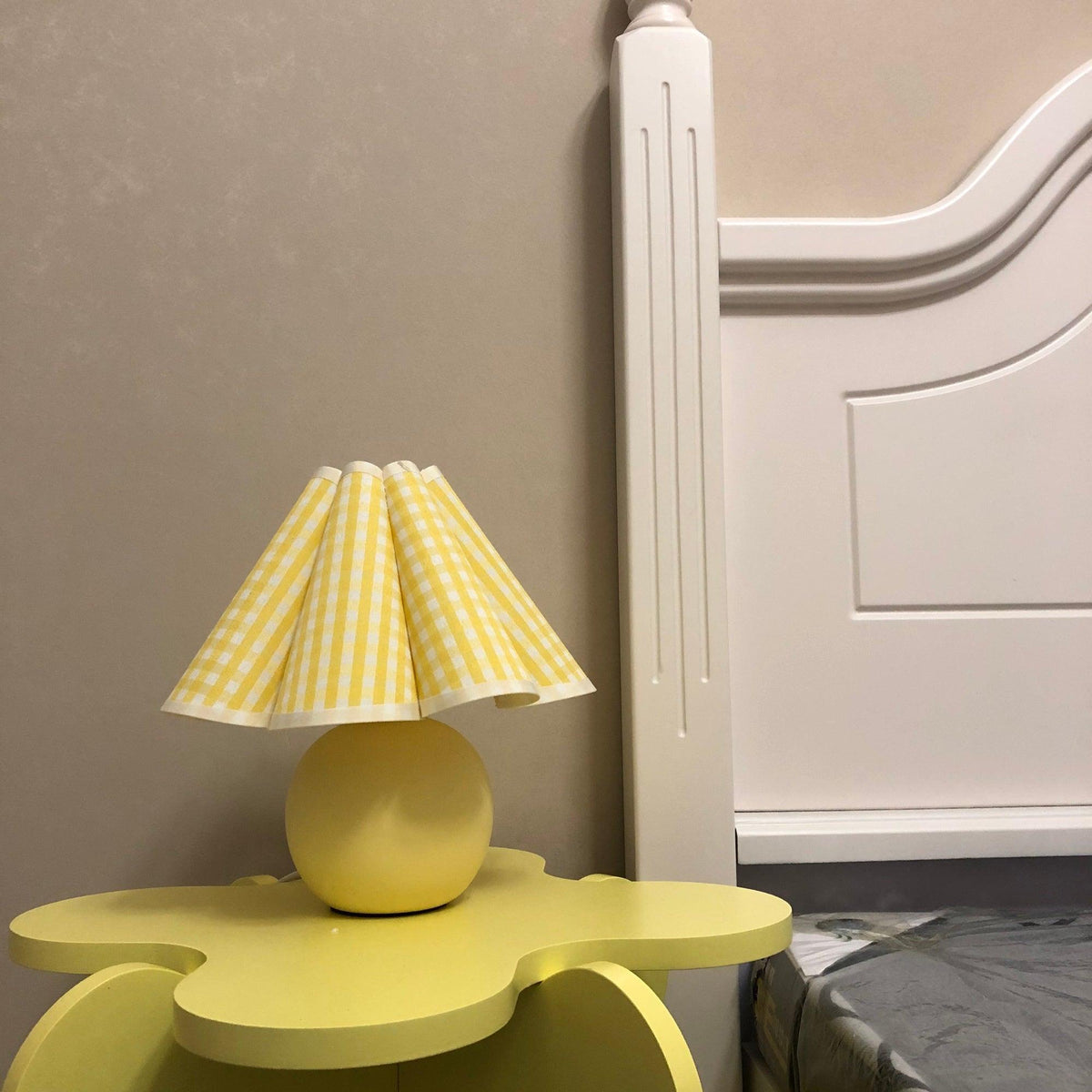 In this photo, a Clérachic table lamp with a yellow plaid pleated shade stands on a cloud-shaped yellow side table next to a white bed frame.