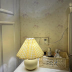 Lit warm, this Clérachic table lamp with a yellow plaid pleated shade stands by a plug socket against floral wallpaper.