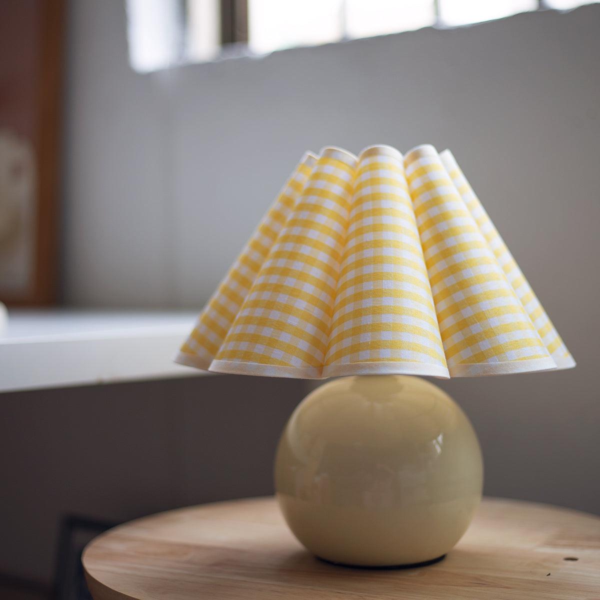 A Clérachic table lamp with a yellow plaid pleated shade and a round pale base, on a wooden stool by a window.