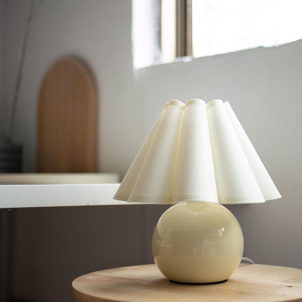 Shown here, a Clérachic table lamp with a cream pleated shade on a round pale yellow base, on a wooden stool by a window.