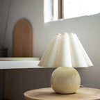 Shown here, a Clérachic table lamp with a cream pleated shade on a round pale yellow base, on a wooden stool by a window.
