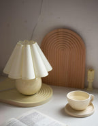This Clérachic table lamp with a cream pleated shade stands in front of a wooden arch, beside a cup and saucer and an open book.