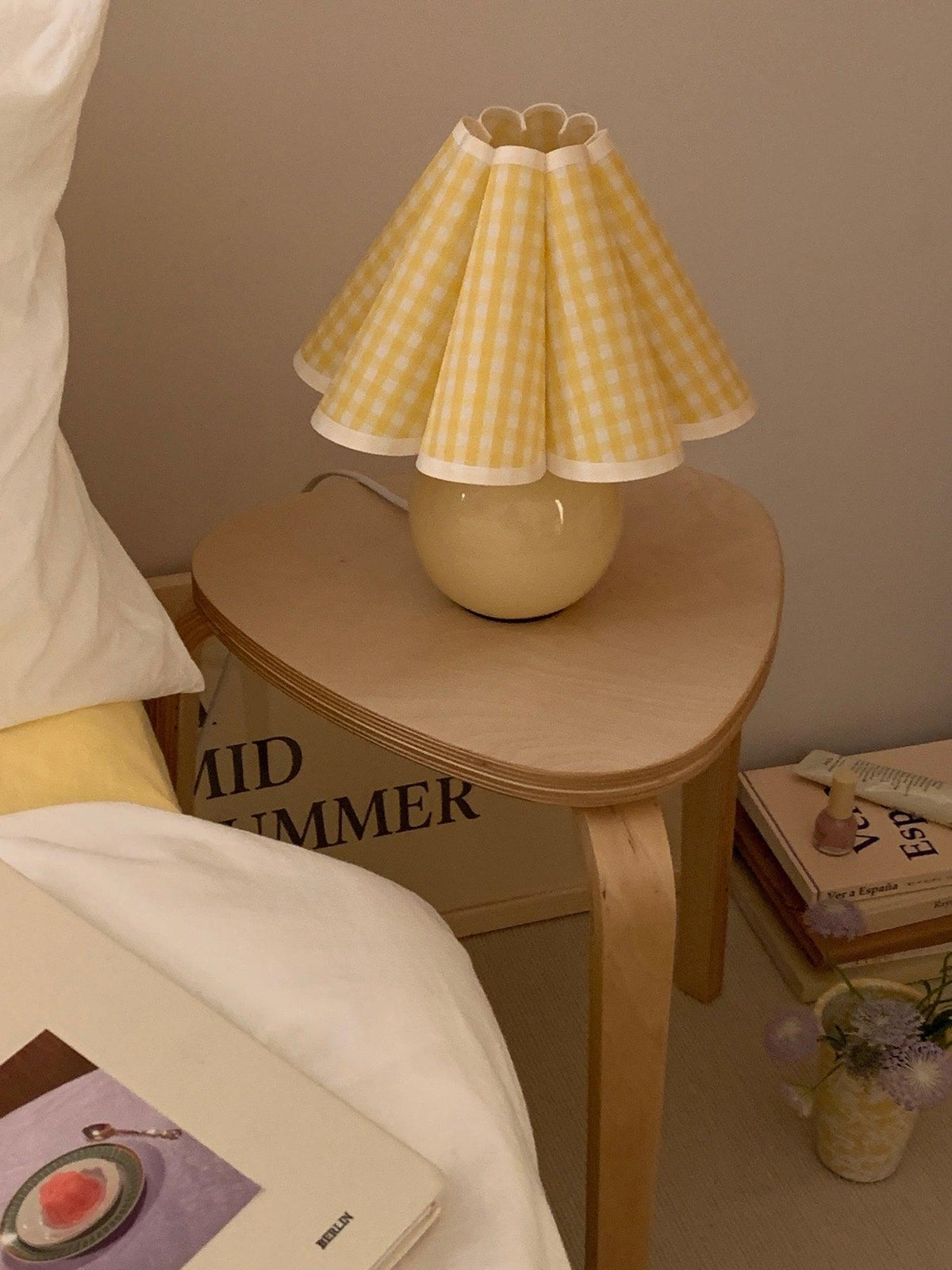 Pictured here, a Clérachic table lamp with a yellow plaid pleated shade stands on a round wooden bedside stool, beside a bed with white linen.