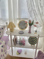A small Clérachic table lamp with a yellow plaid pleated shade sits on a white shelving unit, beside a round mirror and a bedspread.