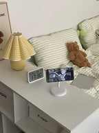 A Clérachic table lamp with a yellow plaid pleated shade stands on a white desk beside a clock, a phone stand and a teddy bear.