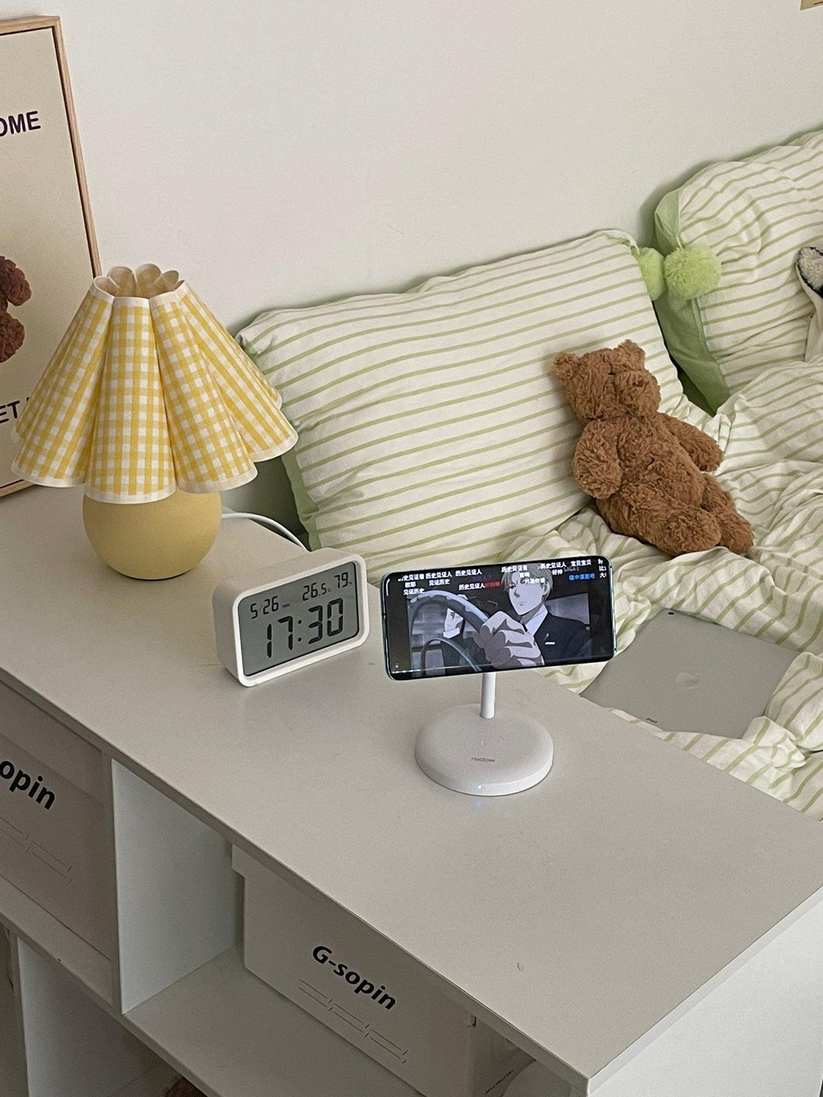 A Clérachic table lamp with a yellow plaid pleated shade stands on a white desk beside a clock, a phone stand and a teddy bear.